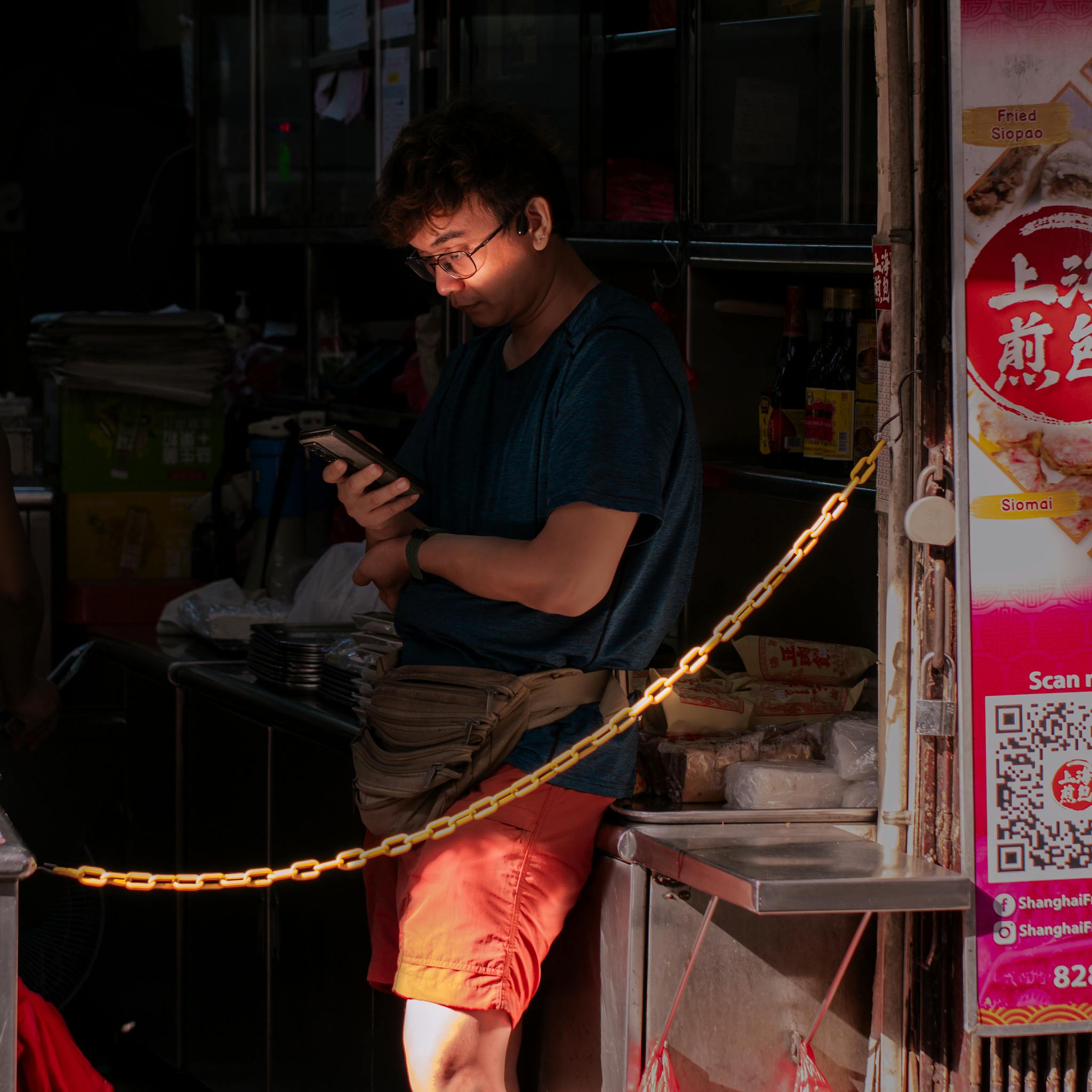 A man stands in a market stall using his phone, illuminated by sunlight.